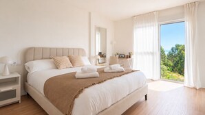 Un dormitorio moderno con una cama grande, mesitas de noche, un escritorio y vistas a zonas verdes a través de un ventanal amplio.