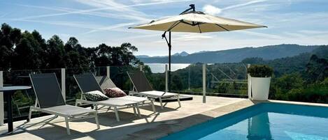 Zona de piscina con sillones de descanso y una sombrilla, con vistas a un paisaje pintoresco.
