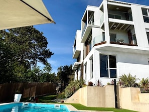 A modern multi-story residential building with a swimming pool, surrounded by greenery and a wooden fence.