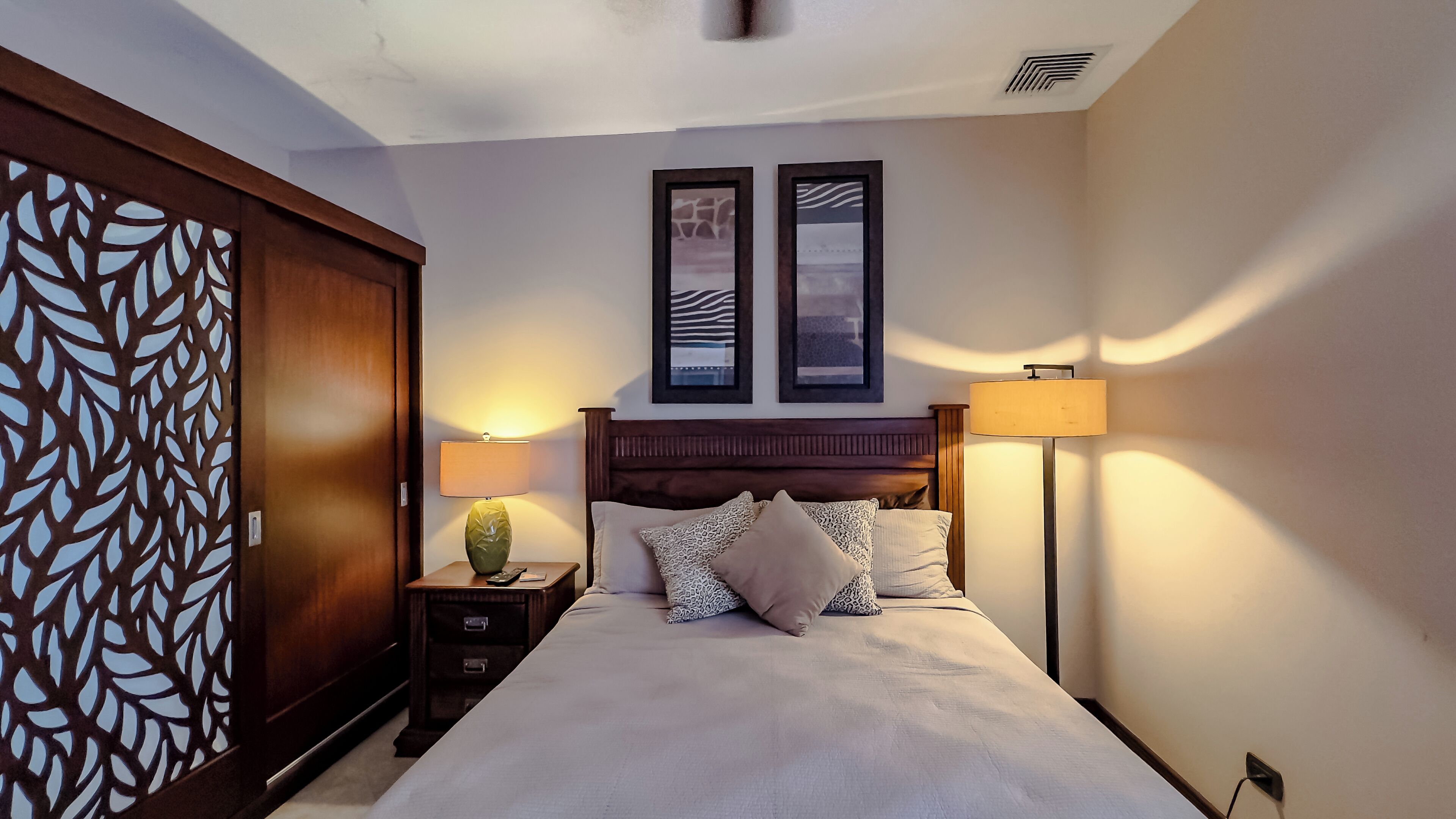 A neatly arranged bedroom with a bed, two bedside tables, a nightstand, a decorative wall panel, and two framed pictures.