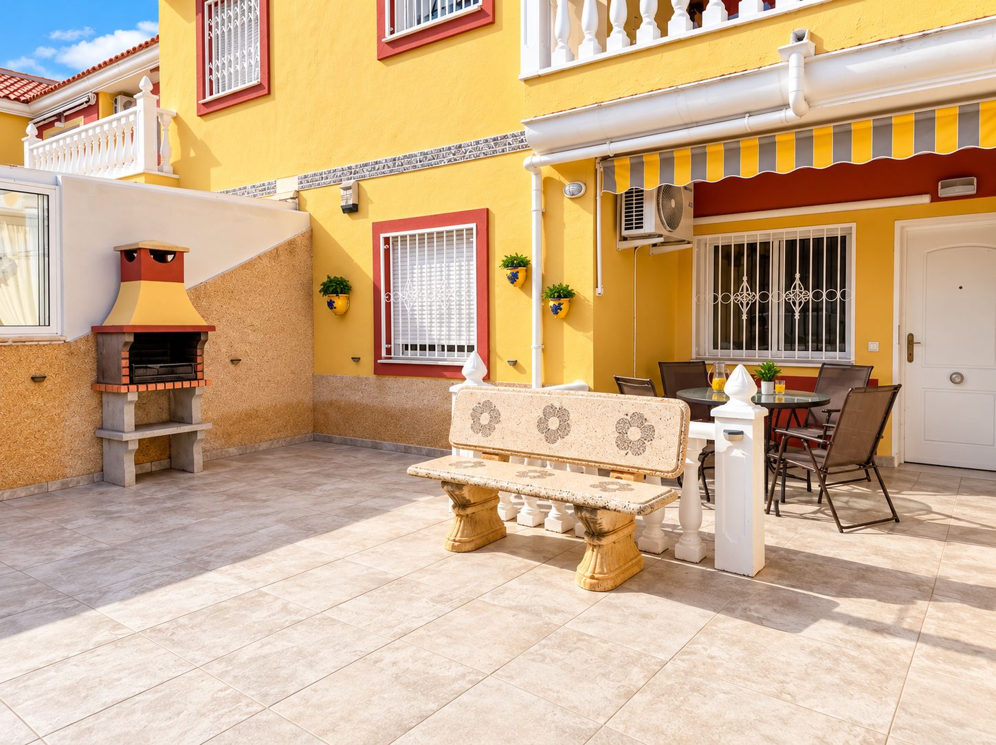 Une terrasse avec un banc en pierre, un barbecue et un espace repas comprenant des chaises et une table.