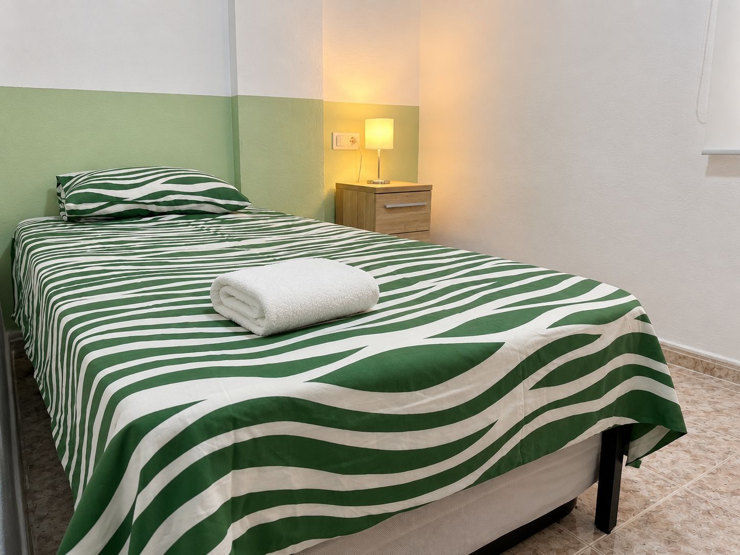 A single-bed room with a green and white striped bedspread, a white towel on the bed, a bedside table with a lamp, and a nightstand.