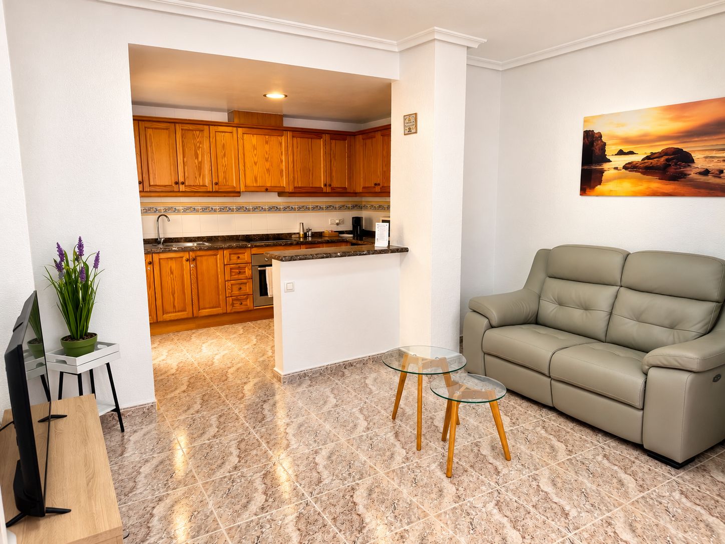 A modern living room with a beige sofa, a glass coffee table, and a kitchen in the background.
