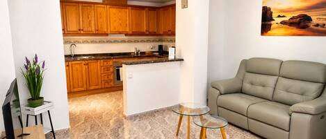A modern living room with a beige sofa, a glass coffee table, and a kitchen in the background.