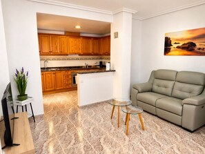 Ein modernes Wohnzimmer mit einem beigefarbenen Sofa, einem Glastisch und einer Küche im Hintergrund.