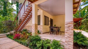 A covered outdoor area with a staircase leading to a second floor, surrounded by lush greenery and a dining table with chairs. - AroCocles Lodge (Cahuita)