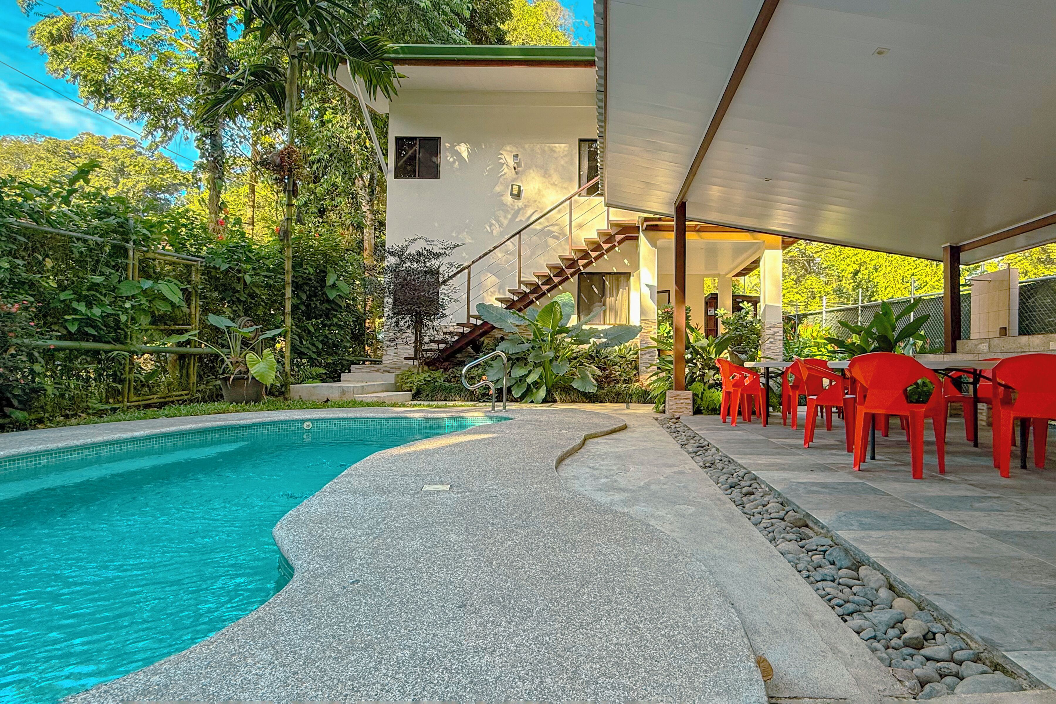 A swimming pool with a stone deck, surrounded by lush greenery and a covered outdoor area with red chairs.