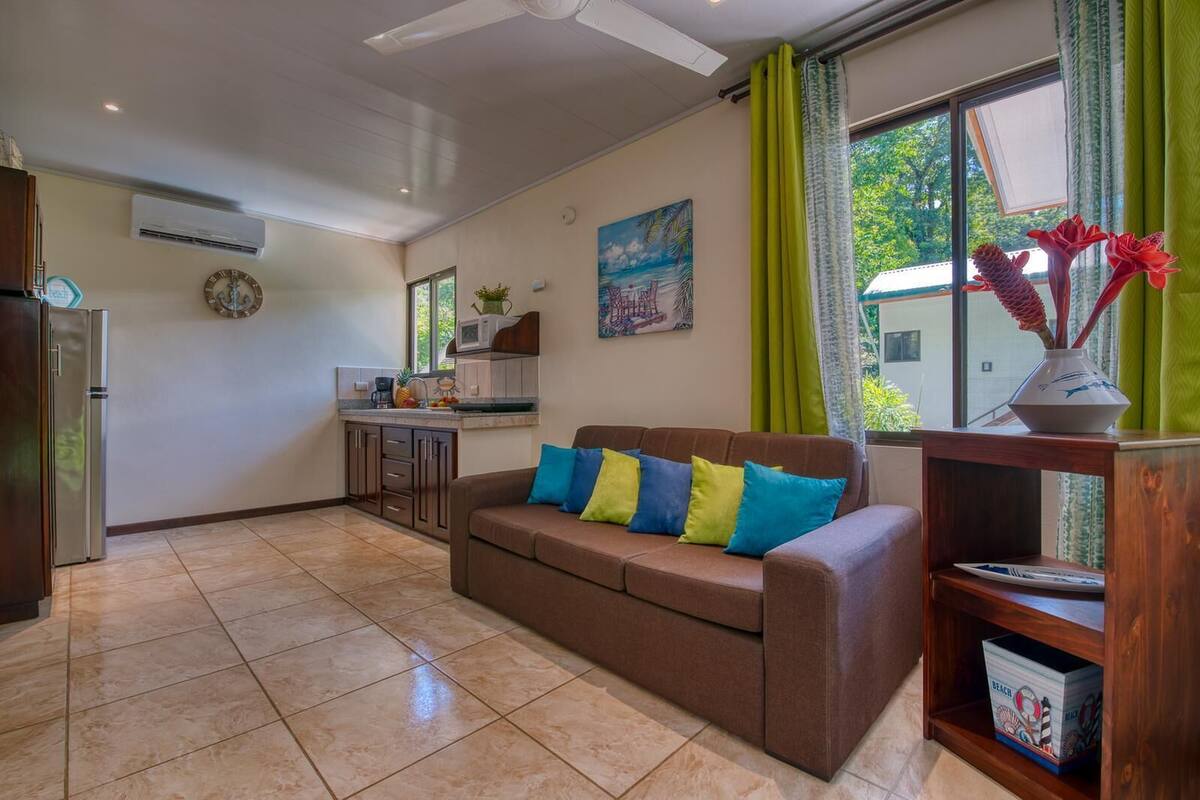A living room with a brown sofa, colorful cushions, a wooden coffee table, and a view of the kitchen and outside through large windows.