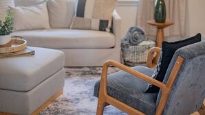 Ein Wohnzimmer mit einer Couch, Hocker und einem Holzstuhl. Eine Lampe steht auf einem Beistelltisch, und ein Fenster ist mit Vorhängen drapiert.