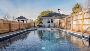 Ein Pool mit Holzterrasse, einem Pavillon und einem Haus mit Schornstein.