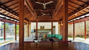 Un espace couvert et spacieux avec un plafond en bois et des colonnes, un coin salon avec un canapé et des coussins, et une vue sur une cuisine et un espace extérieur.