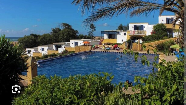 Una piscina rodeada de edificios modernos con balcones, cielo despejado y vista al mar.