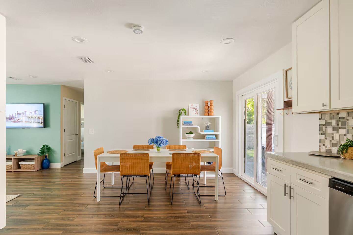 Una cocina moderna con gabinetes blancos, un comedor con una mesa blanca y sillas de mimbre, y un amplio ventanal que proporciona luz natural.