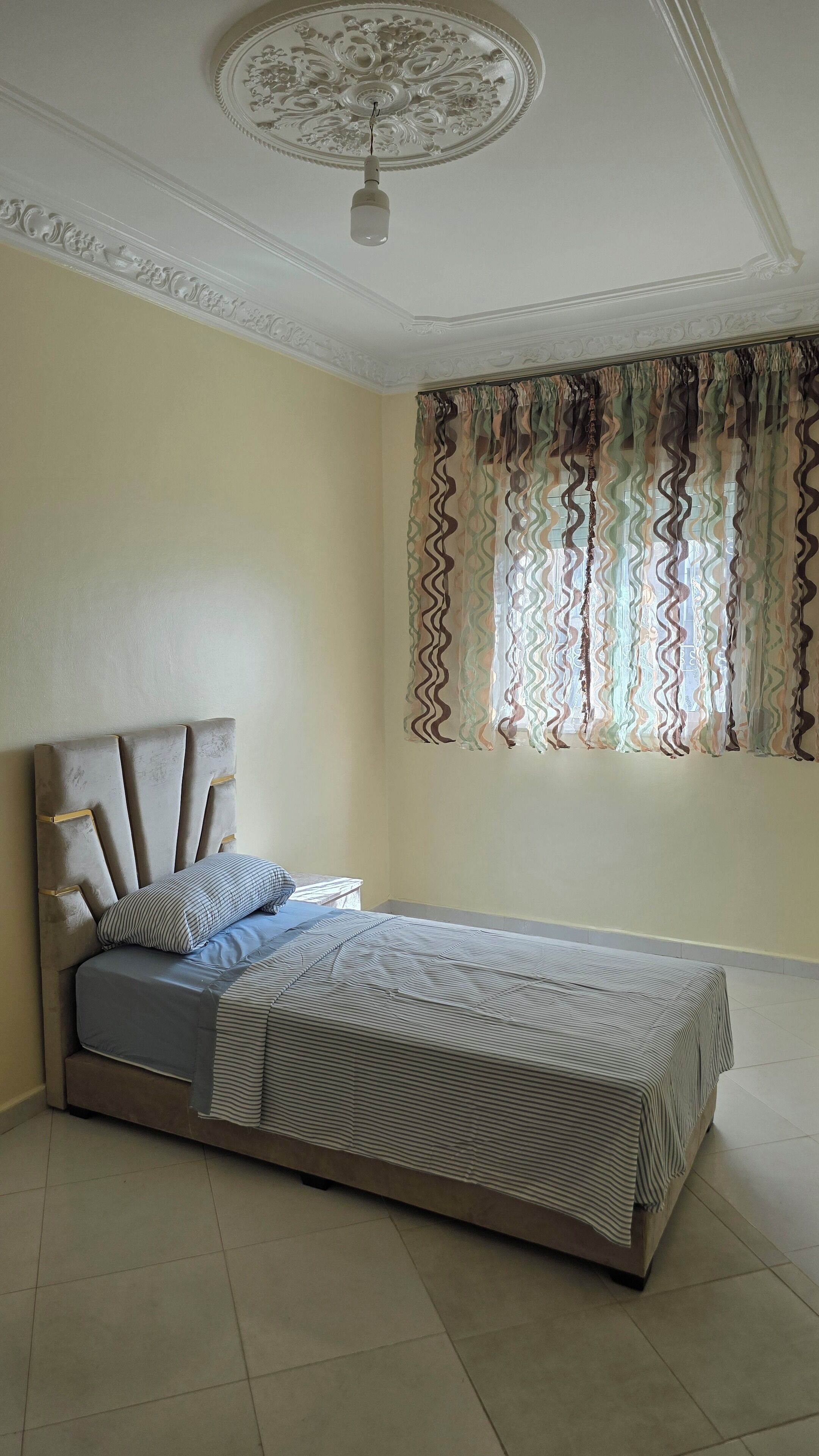 Une chambre à coucher avec un lit, un luminaire au plafond et une fenêtre ornée de rideaux à motifs.