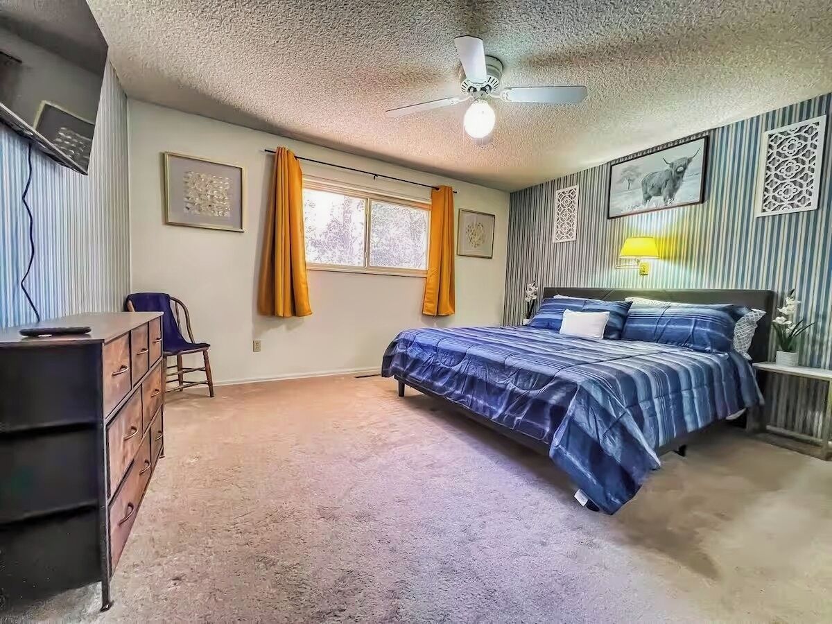 Een slaapkamer met een bed, een dressoir, stoel, raam, gordijnen en een plafondventilator.