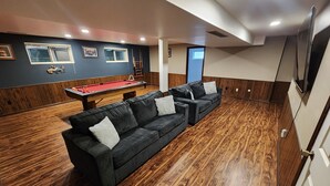 Una habitación con una mesa de billar, dos sofás, suelo de madera y un televisor montado en la pared.