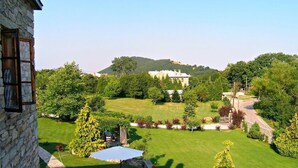 A stone building with a view of a well-maintained lawn, two white umbrellas, and a distant hill with a house. - La Moara (Amyntaio)