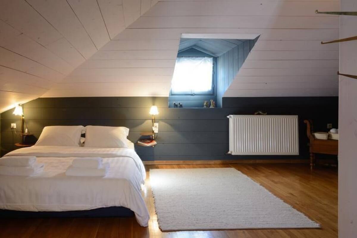 Quarto com cama, duas luminárias de cabeceira, uma janela, radiador e piso de madeira.