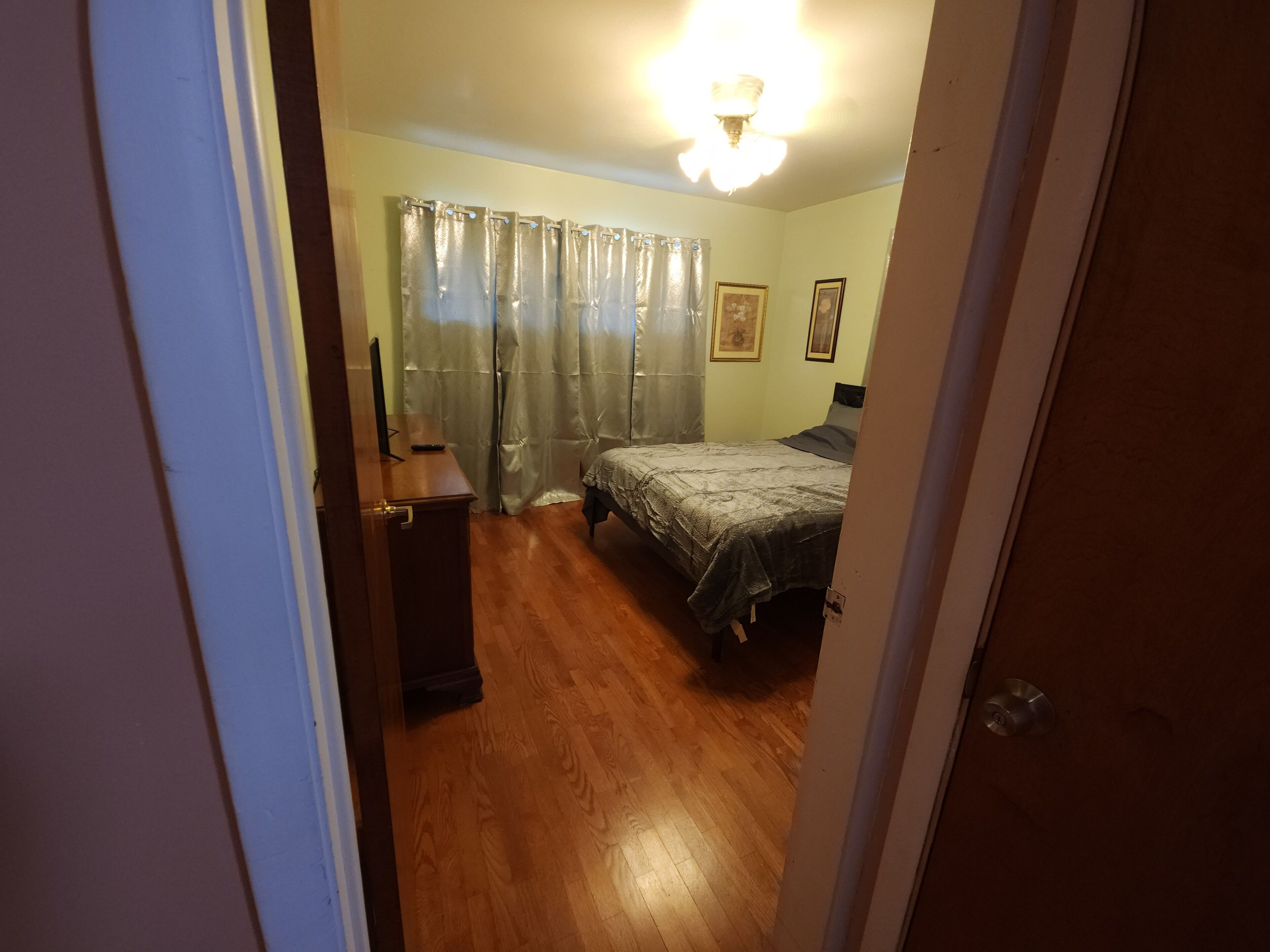 Une chambre à coucher avec un lit, un bureau et une chaise. Il y a une fenêtre avec des rideaux et un luminaire au plafond.