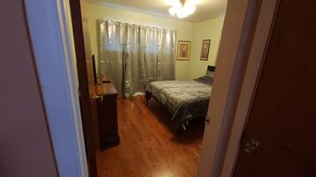 Une chambre à coucher avec un lit, un bureau et une chaise. Il y a une fenêtre avec des rideaux et un luminaire au plafond.