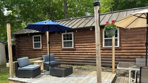 A wooden deck with outdoor furniture, a blue umbrella, and a hanging planter with flowers. - Cabin with big fenced yard-firepit-walking distance to Sacandaga lake- pets ok (Broadalbin)