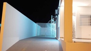 Un couloir étroit et bien éclairé, avec une grille métallique au bout, qui mène à un bâtiment à la façade blanche.