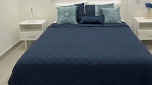 Un lit bien fait, avec une couverture bleu foncé, une tête de lit blanche et des tables de chevet assorties, chacune surmontée d’une petite table.