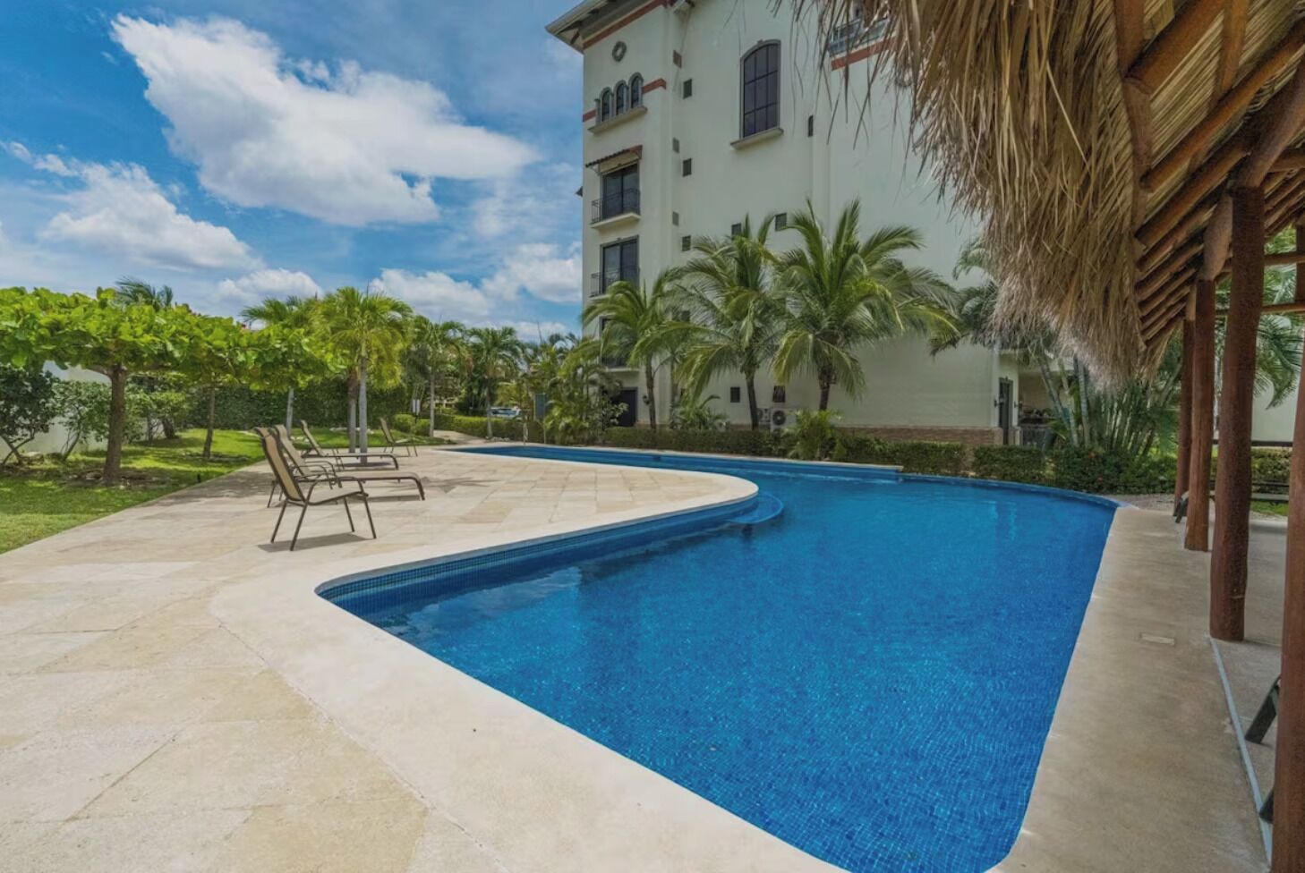 A swimming pool with lounge chairs, surrounded by palm trees and a building with multiple windows.