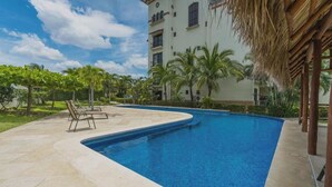 A swimming pool with lounge chairs, surrounded by palm trees and a building with multiple windows. - Stunning Oceanview Condo Steps to Playa Flamingo w/ Pool, BBQ & Beach Access (Playa Flamingo)