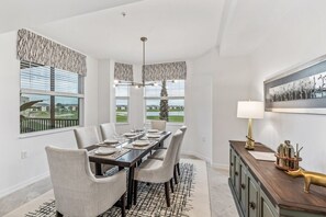 Comedor con una gran mesa dispuesta para seis comensales, ventanas amplias con persianas, un candelabro y un aparador con lámpara y objetos decorativos.