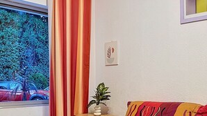A cozy room with a sofa, a small wooden side table, a potted plant, and a view of trees and cars outside. - Studio Cannes, 350 m from the sea (Cannes)