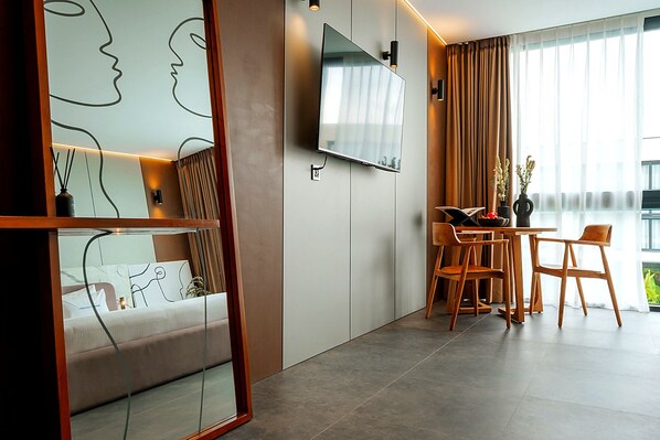 A modern room with a bed, a TV mounted on the wall, a dining area with chairs, and a large window with curtains.