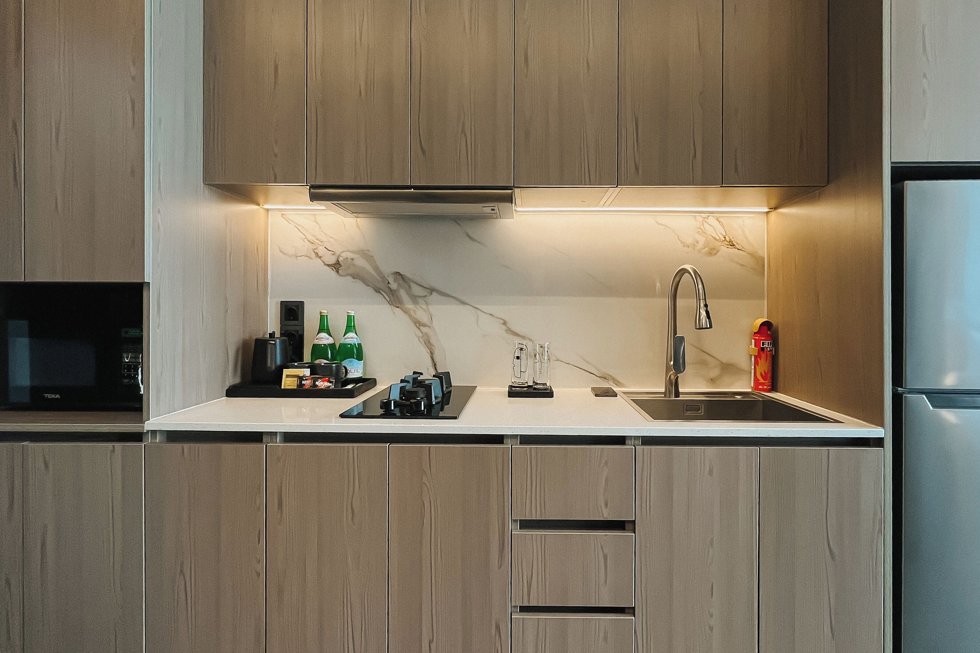 A modern kitchen with wooden cabinets, a marble backsplash, a stainless steel refrigerator, and a microwave.