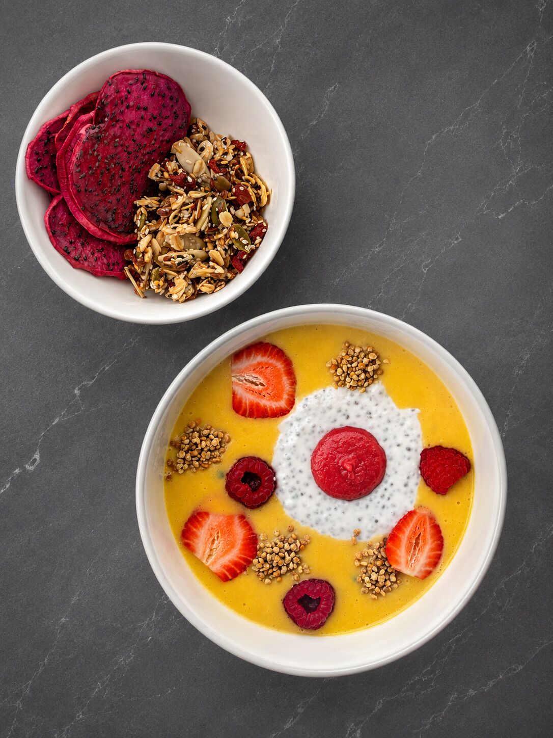 Two bowls of food, one with dragon fruit and granola, the other with a creamy dish topped with berries and seeds.