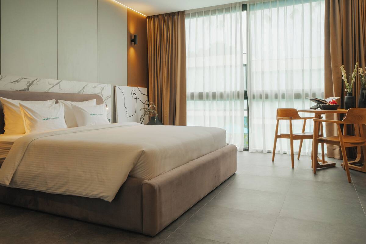 A modern hotel room with a large bed, a desk with chairs, and a view of the city through sheer curtains.