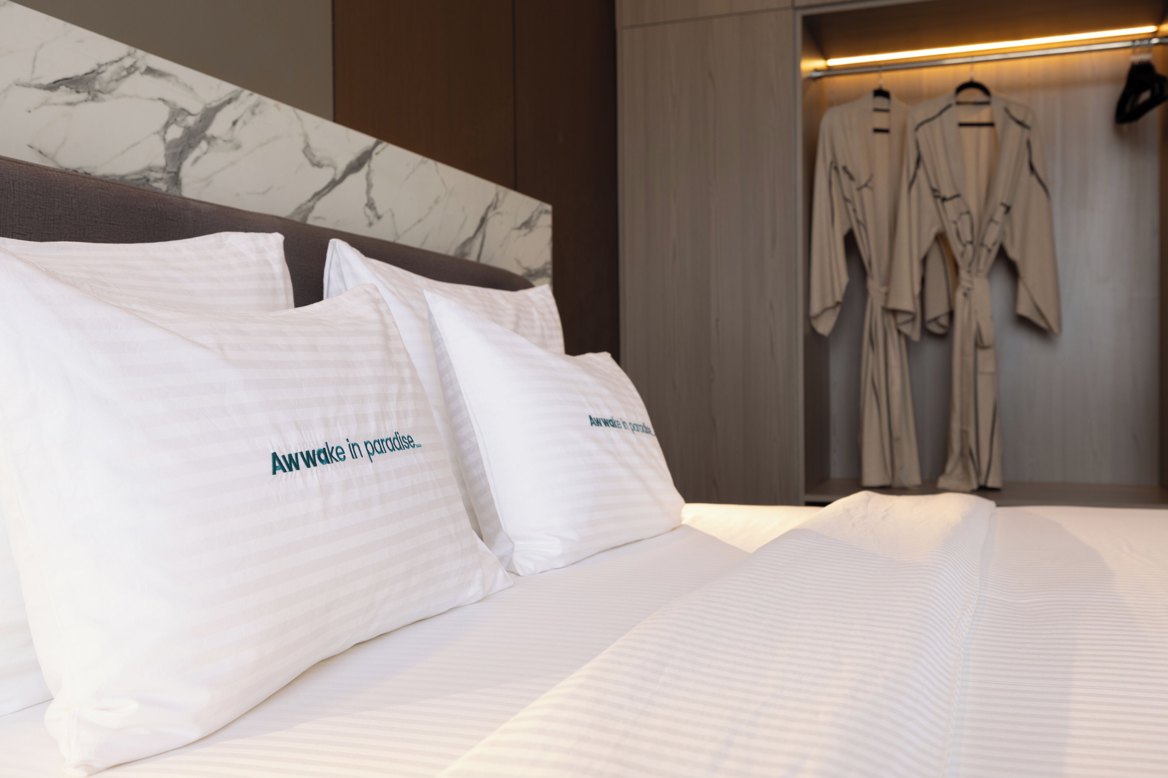 A hotel room with a neatly made bed, two pillows with text, and a robe rack with robes.