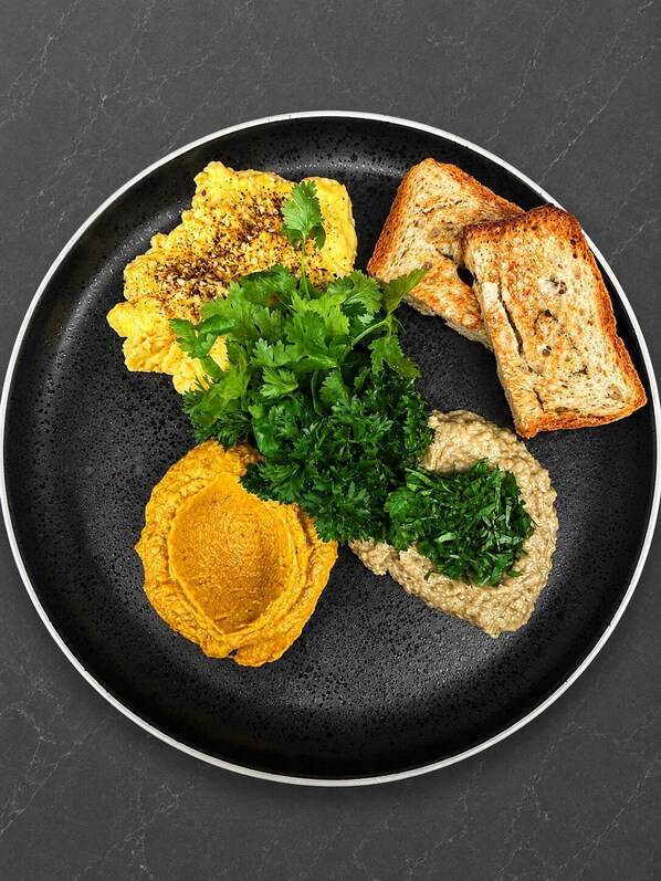 Une assiette noire contenant des œufs brouillés, des toasts et deux sortes de tartinades, le tout garni de persil.