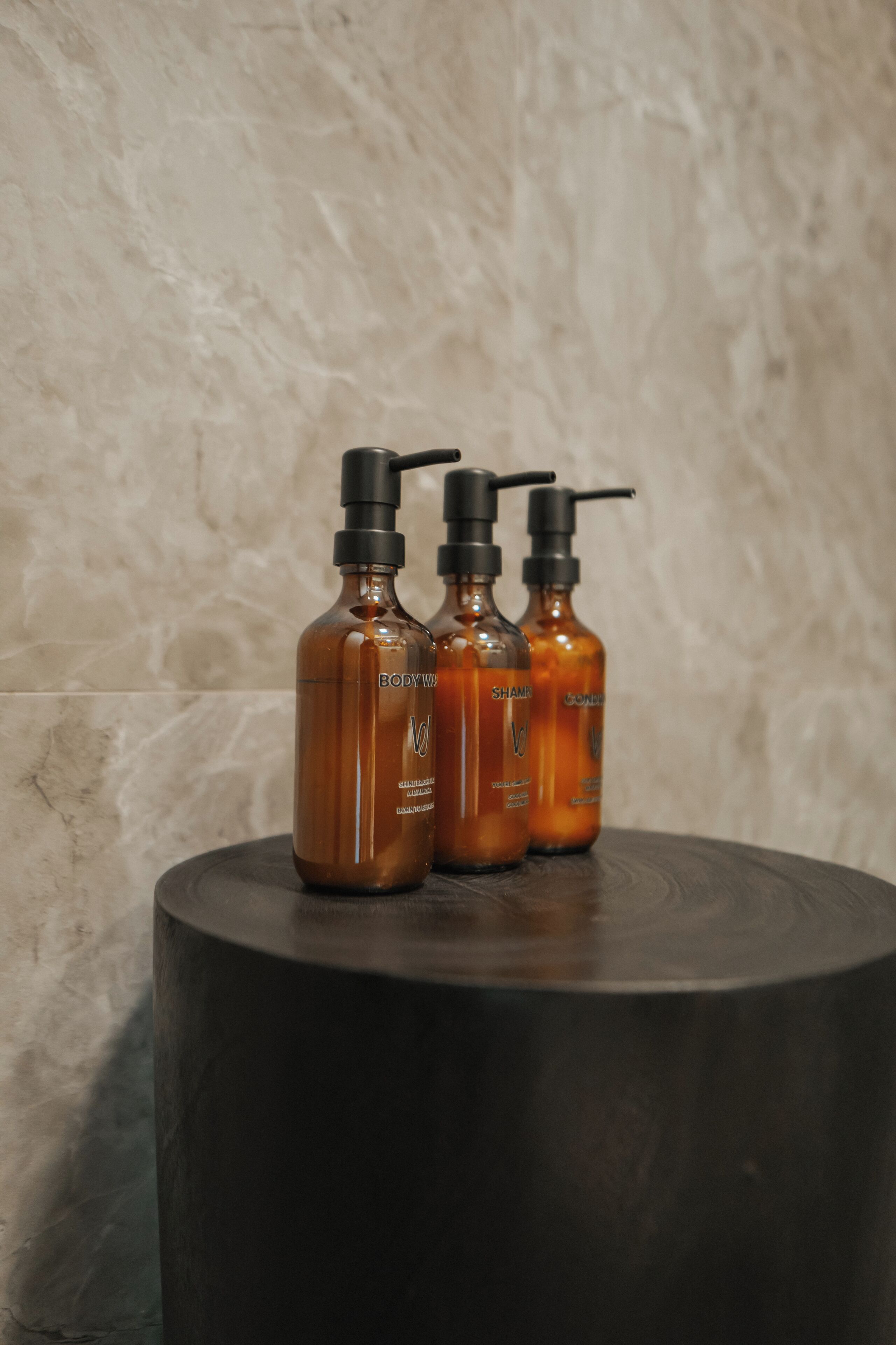 Three amber bottles with black pumps on a round black table against a marble wall.