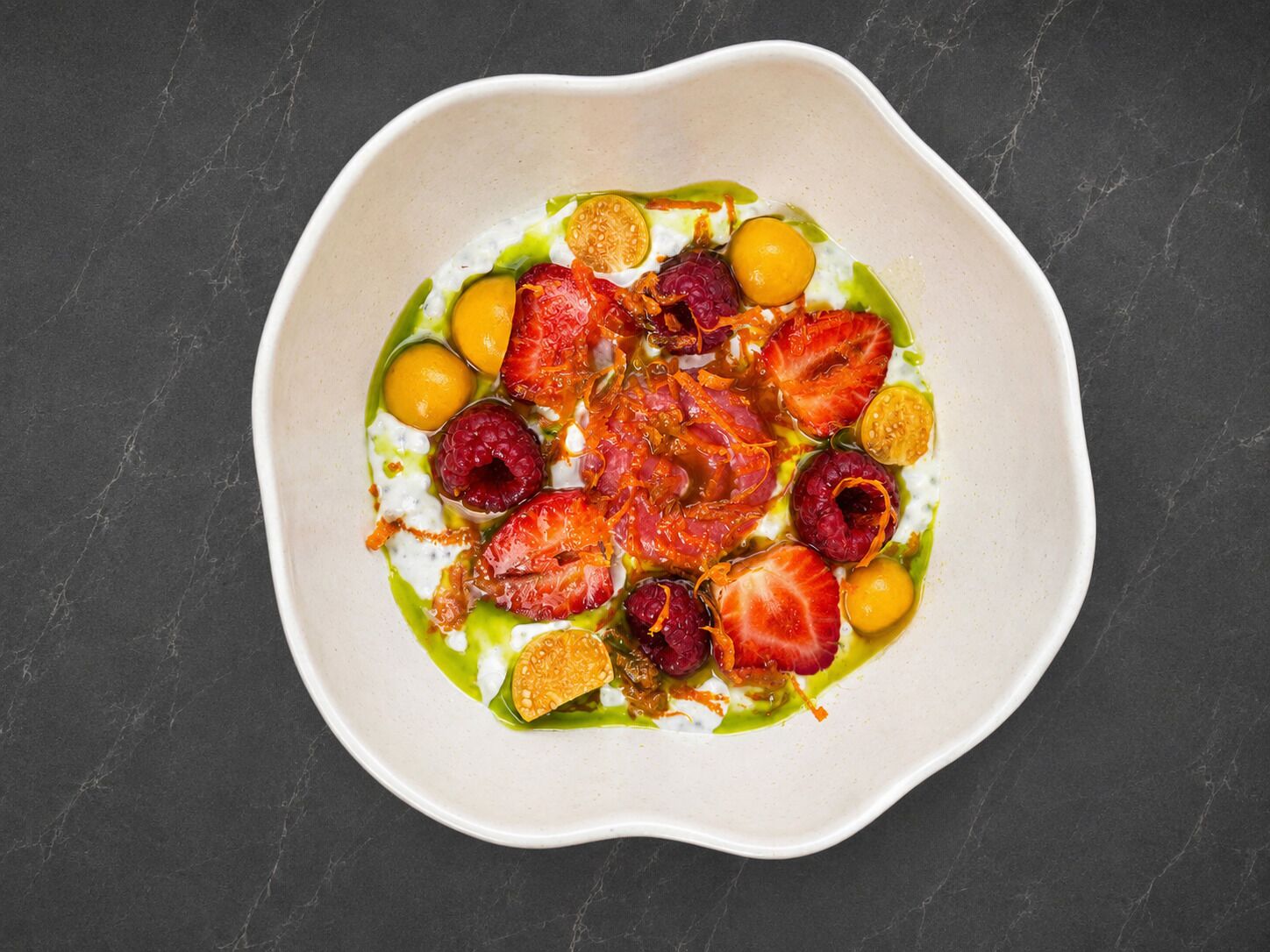 A white bowl with a variety of fruits and a green sauce on a dark surface.