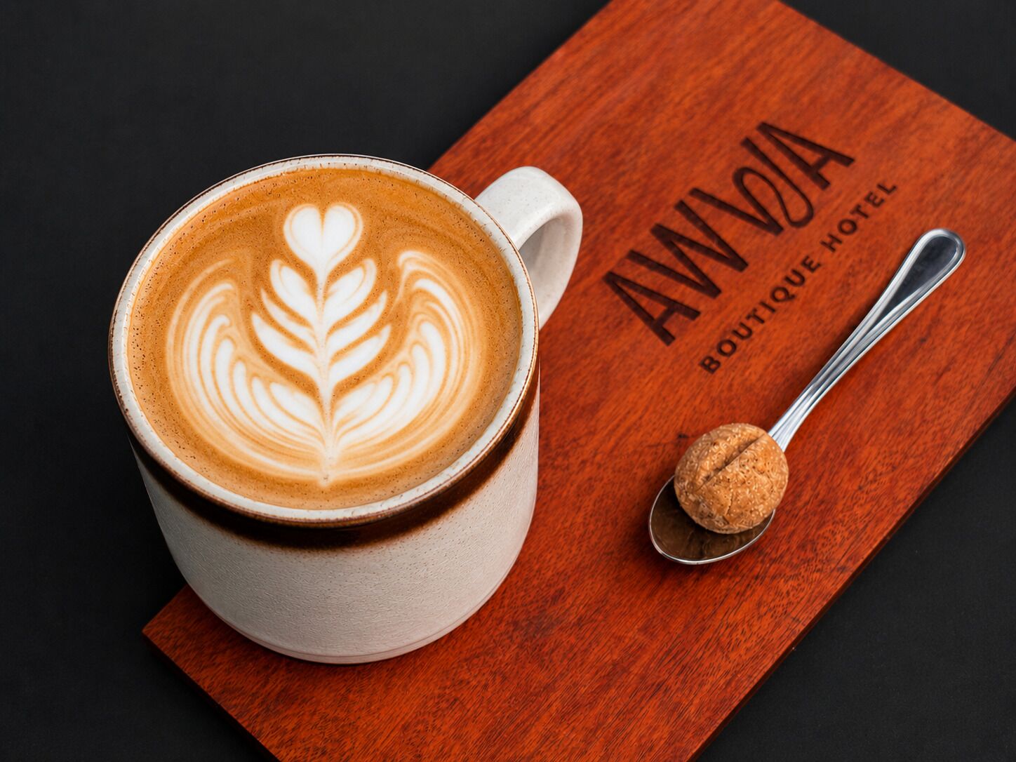 Eine Tasse Kaffee mit Latte-Art in einer weißen Tasse, ein Löffel und ein Zuckerpaket, sowie ein Untersetzer aus Holz mit dem Text 'AWVIA BOUTIQUE HOTEL'.