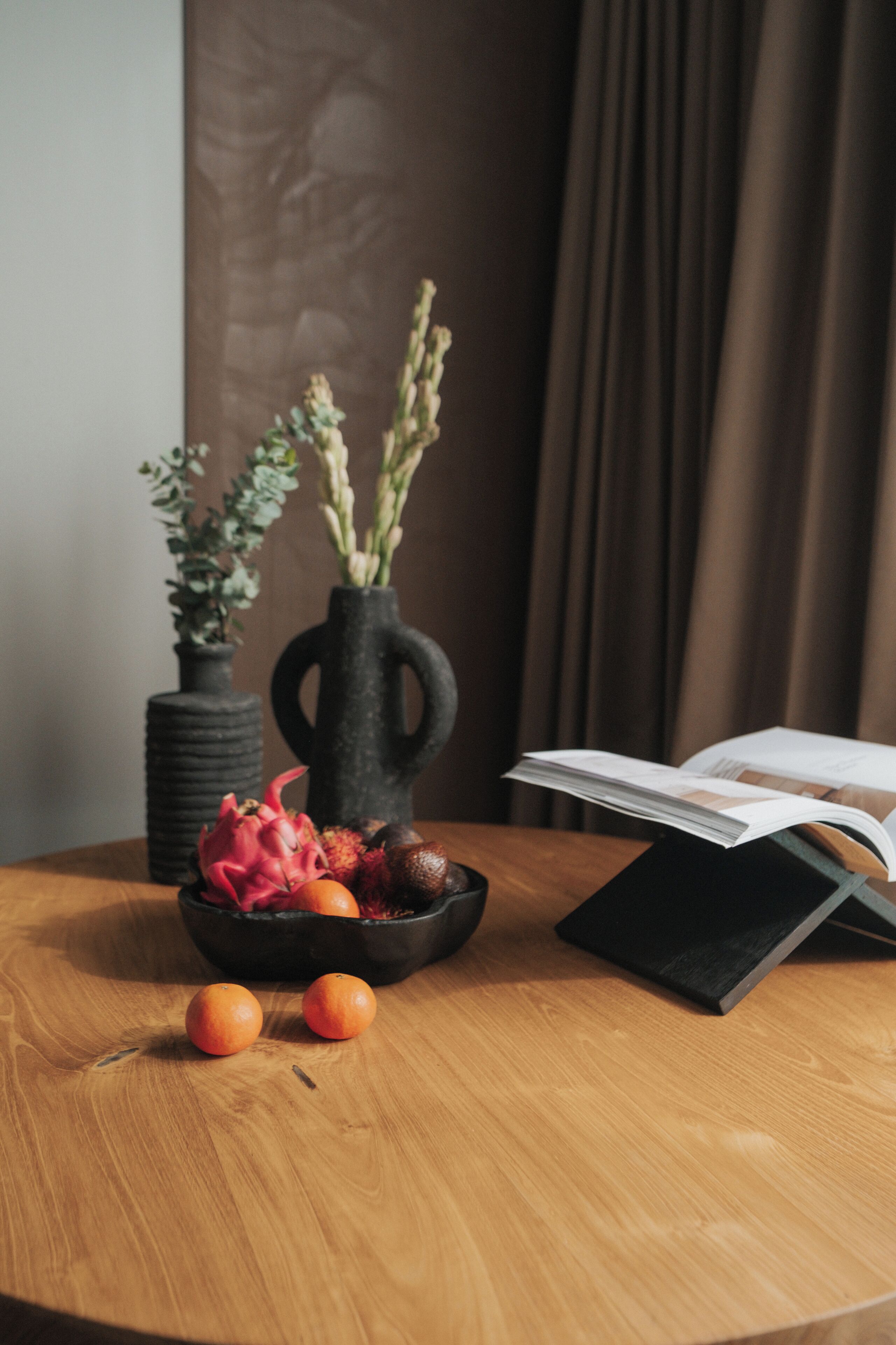 A round wooden table with a black vase containing dried flowers, a bowl with fruits, and a book.