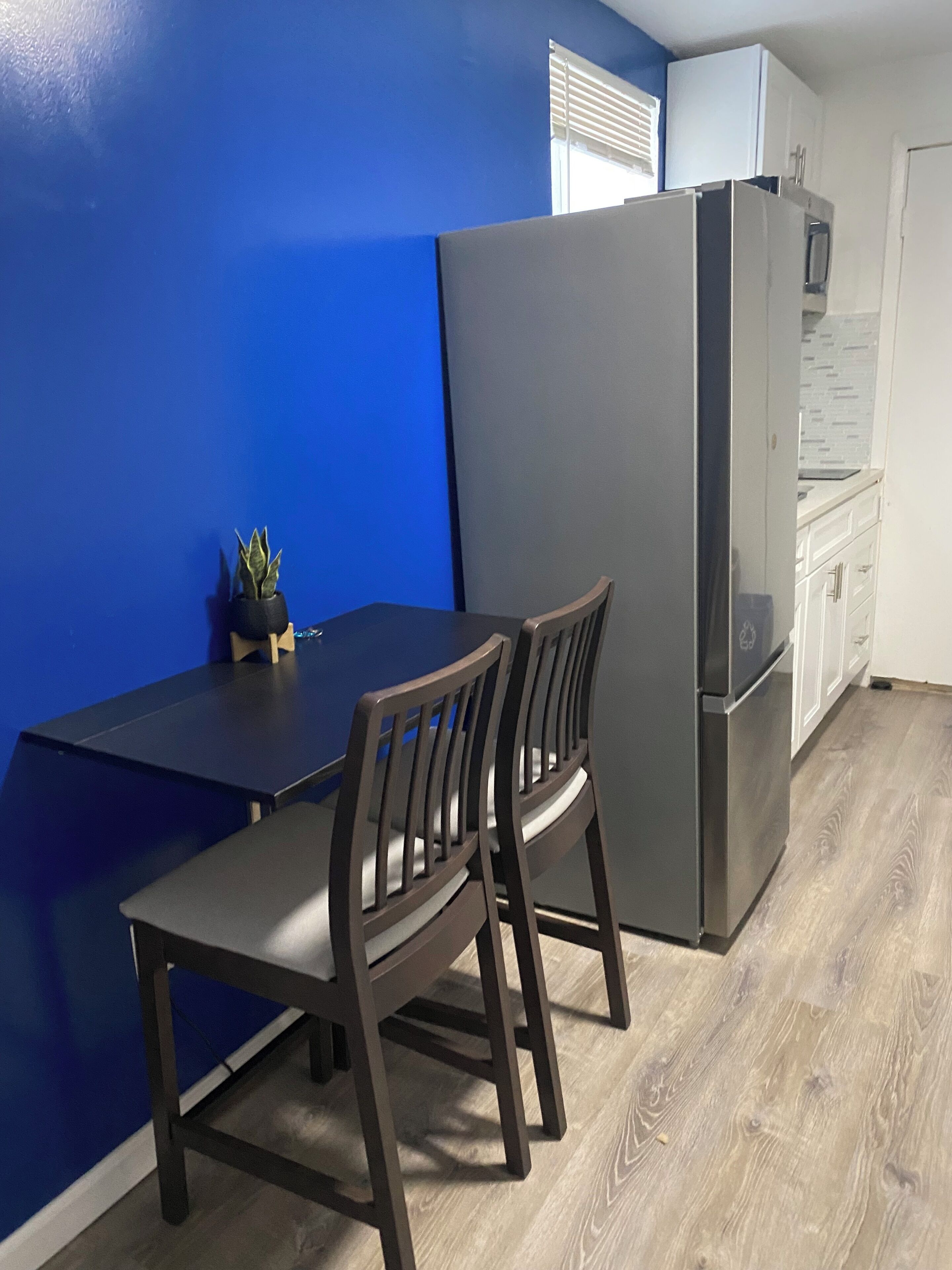 Une cuisine de taille réduite avec un mur d’accent bleu, une table en bois foncé, deux chaises, un réfrigérateur et un four à micro-ondes.