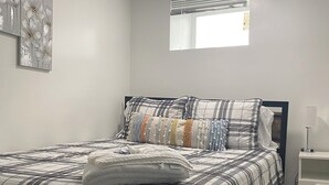Un lit bien fait, recouvert d’une couverture à carreaux, une table de chevet avec une lampe et une fenêtre avec des stores.