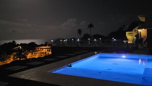 Una piscina con una persona en el agua, rodeada de una zona de solárium con vistas al mar y un edificio al fondo.
