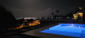 A swimming pool with a person in the water, surrounded by a deck area with a view of the sea and a building in the background. - Private, Cozy 1-bedroom apartment with unobstructed views in serene Patillas (Patillas)