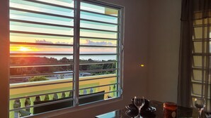 Ein Zimmer mit großem Fenster und Blick auf eine malerische Landschaft, inklusive Sonnenuntergang über Wasser und Grünflächen.