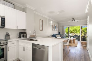 A modern kitchen with white cabinetry, a built-in dishwasher, and a microwave. There is a dining area with a view of the outdoors. - Blue Waters Stay - Kayaks - 2BR 2.5BA Coastal Villa (Duck Key)