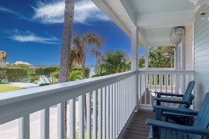Een overdekt terras met witte reling, twee blauwe Adirondack-stoelen en uitzicht op een strand en palmbomen.