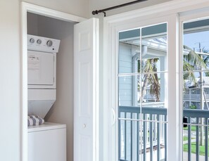 Une buanderie équipée d’une machine à laver et d’un sèche-linge superposés, une porte donnant sur un balcon avec vue sur des palmiers et une maison.