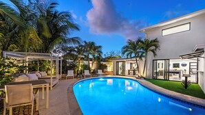 A swimming pool with lounge chairs and a covered seating area, surrounded by palm trees and a modern building. - Villa Azur 8BR Outdoor Oasis with Poolside Bar (Miami)